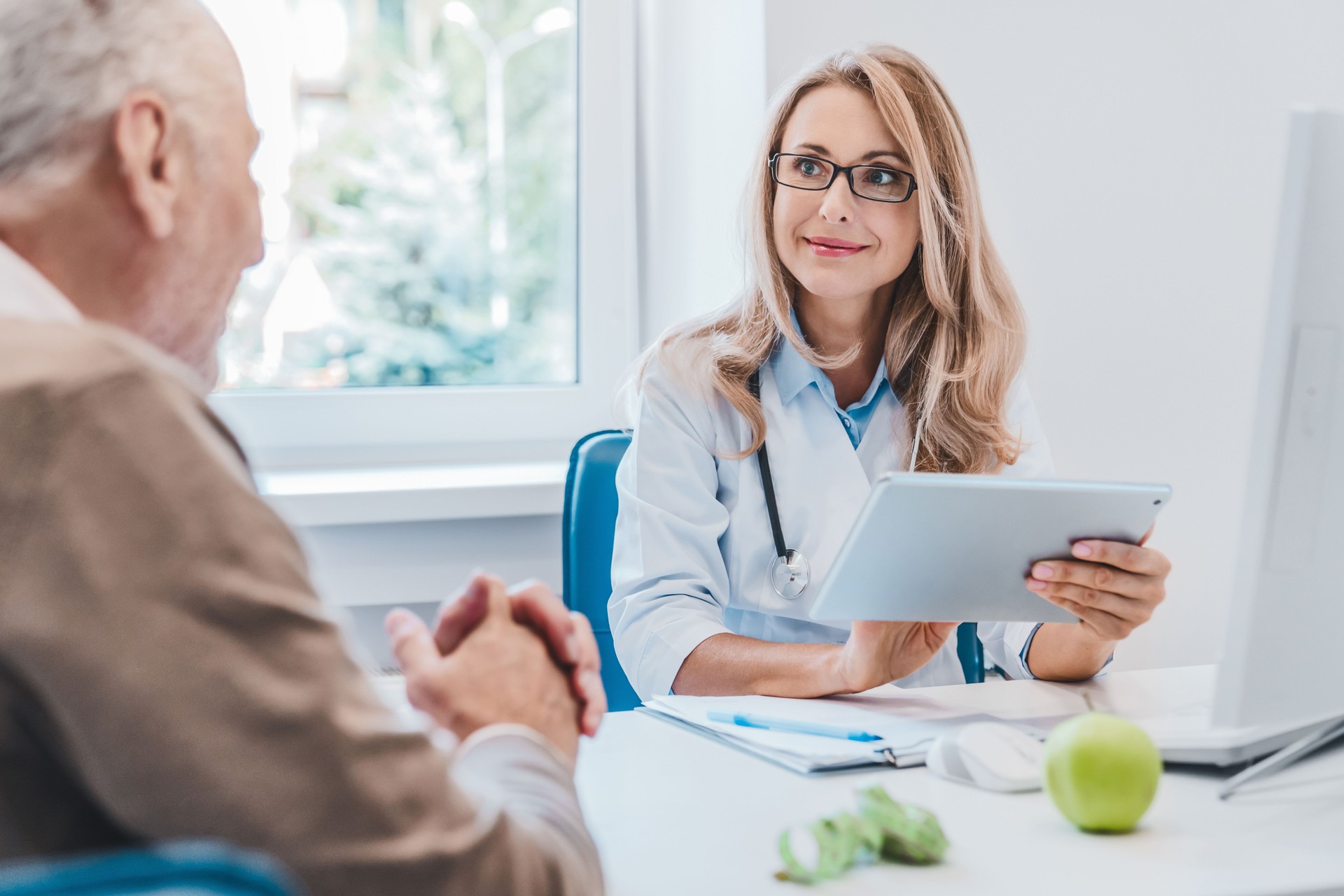 Middle aged female doctor nutritionist in white medical coat consulting her old patient using digital tablet Middle aged female doctor nutritionist in white medical coat consulting her old patient using digital tablet